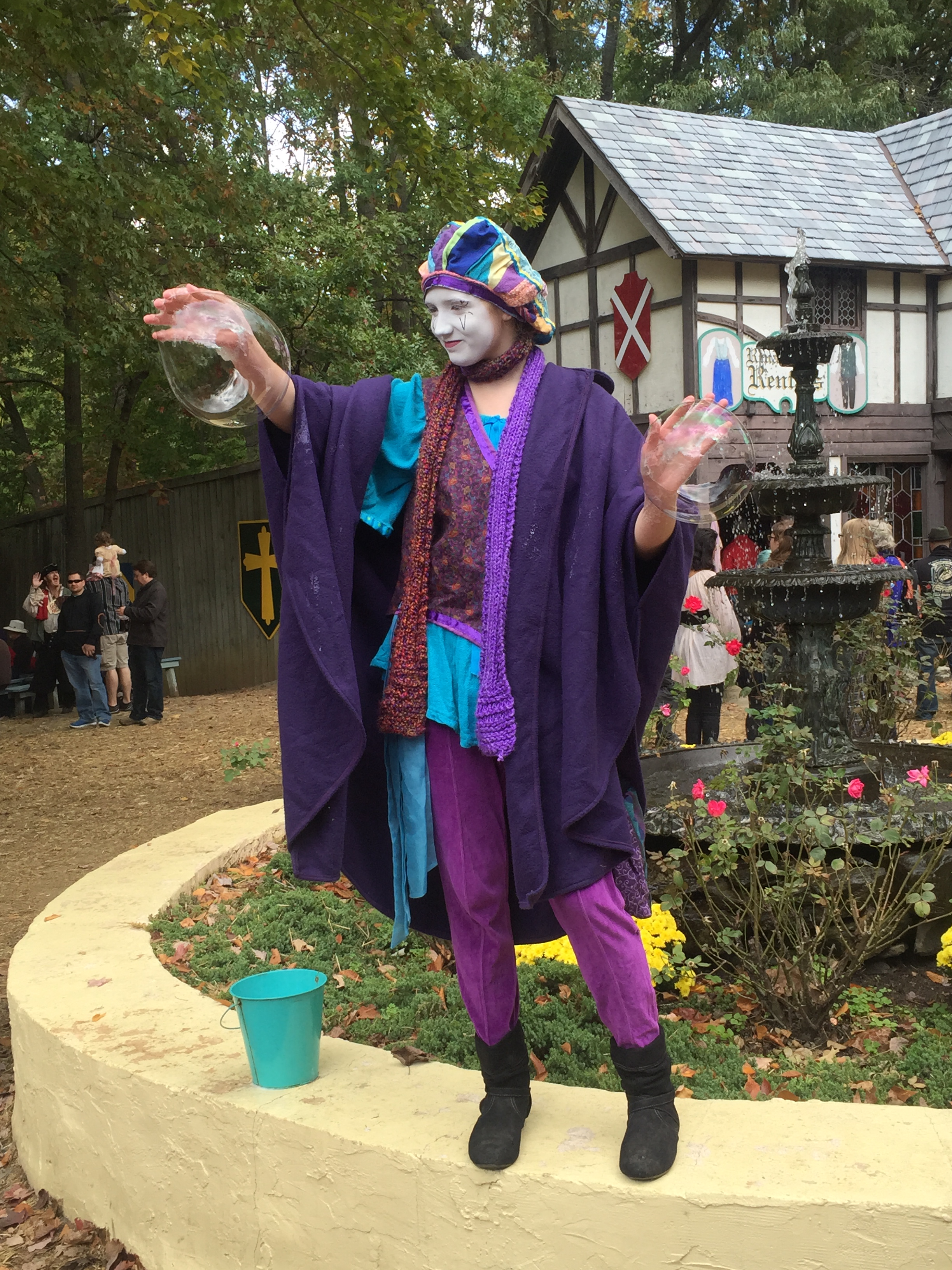 Jester dressed in multi-colored clothing blowing bubbles using only her hands